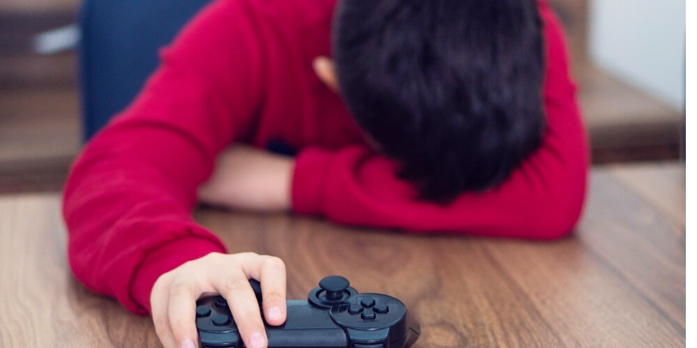 Niño con camisa roja de manga larga recostado en una mesa de madera con la cabeza en su brazo isquierdo escondiendo la cara. Su mano derecha aguanta un control de videojuegos.