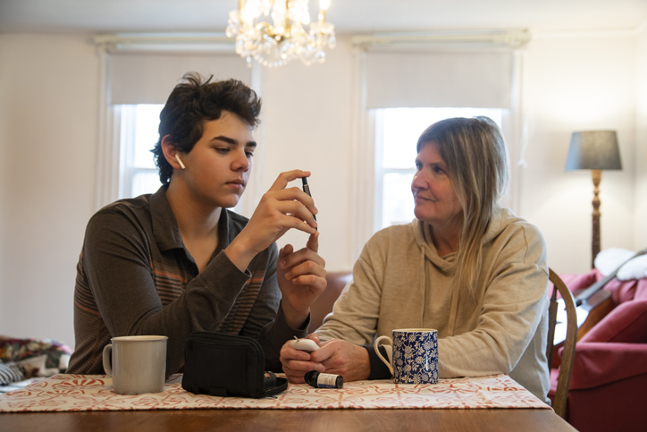 A mother supporting her teenaged diabetic son at home while he tests his blood sugar levels. Photographed in North America.